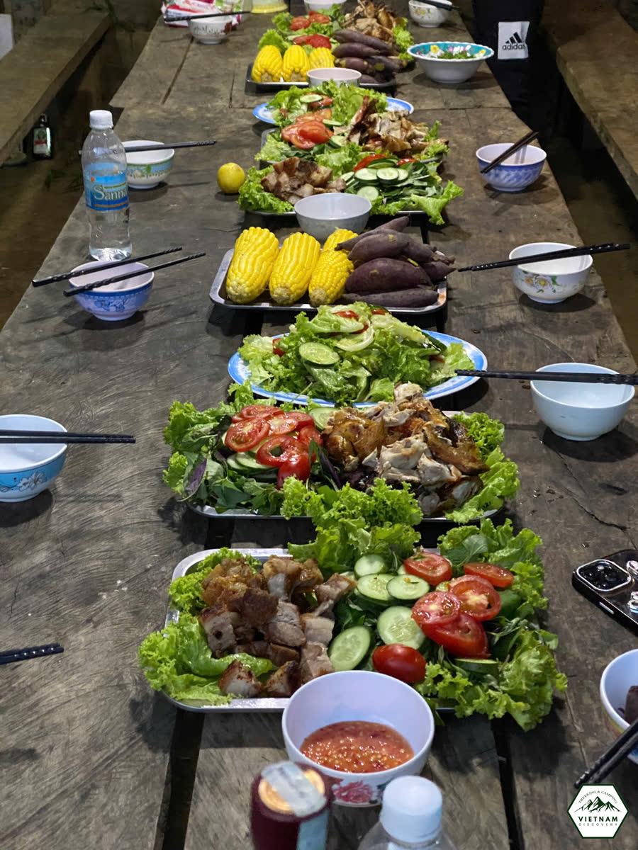 Bữa ăn của đoàn bày trên bàn gỗ dài, nhiều rau, bắp và món mặn