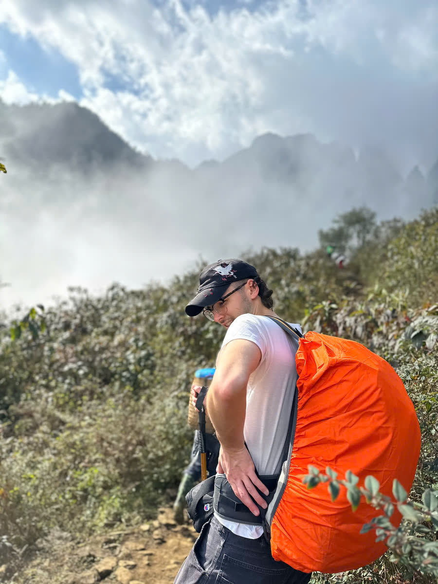 Người đi bộ đeo balô bọc áo mưa màu cam trên đường lên Ngũ Chỉ Sơn, phía sau là mây và núi đá